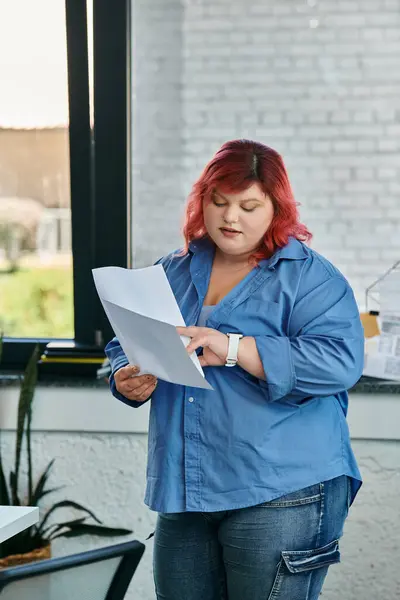 A plus size woman with bright red hair reads a document in a modern office. — Stock Photo