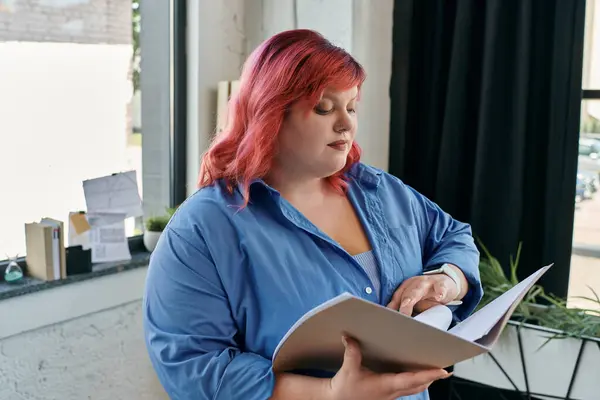 Una donna plus size in camicia blu e capelli rosa legge un documento in un ufficio moderno. — Stock Photo