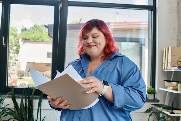 A plus size woman with pink hair, dressed in a blue shirt, smiles as she reviews documents in her office. — Stock Photo