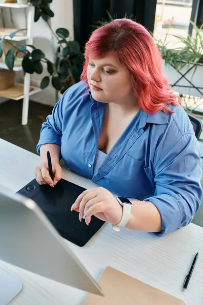 Una mujer de talla grande con cabello rosa trabaja en una tableta de dibujo digital en un estudio brillante. - foto de stock