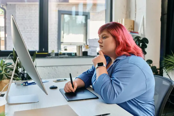 Una mujer de talla grande con el pelo rosado vibrante se está concentrando en su trabajo mientras se dibuja en una tableta digital. - foto de stock