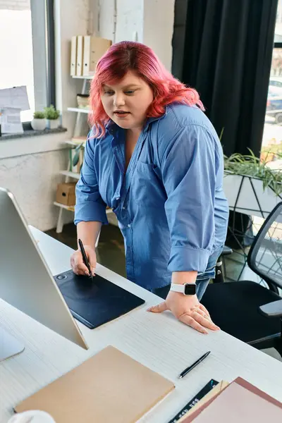 Una mujer de talla grande con pelo rosa brillante y camisa azul utiliza un lápiz óptico en una tableta de dibujo digital. - foto de stock