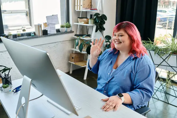 A plus size woman with pink hair, smiles and waves while video chatting from her modern office. — Stock Photo