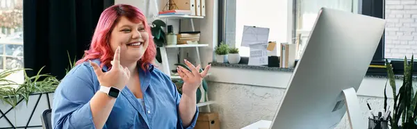 A plus size woman in a blue shirt smiles and gestures, sitting in front of a computer. — Stock Photo