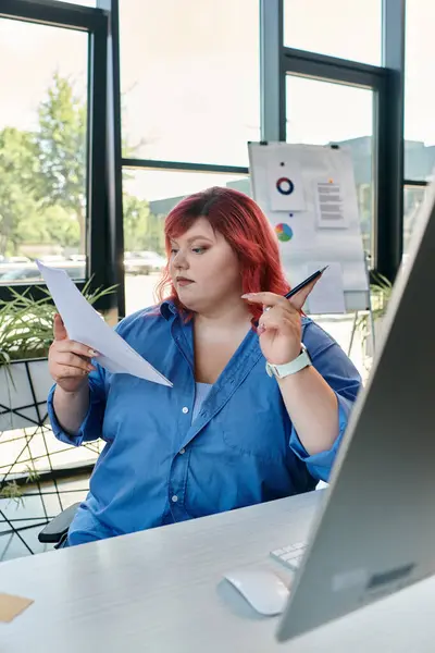 Женщина plus size сидит за столом в современном офисе, рассматривая документы, держа в руках ручку. — стоковое фото