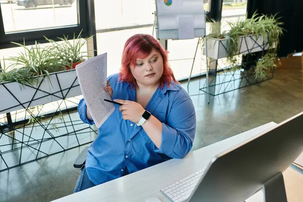 A plus size woman points to a document while sitting at a desk in front of a computer. — Stock Photo