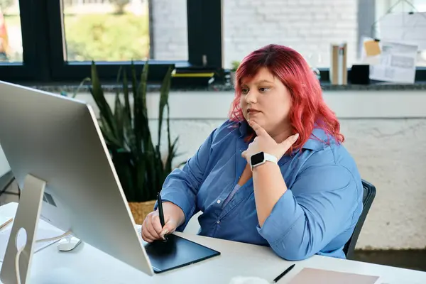 Una mujer de talla grande, con el pelo rojo vibrante, utiliza una tableta de dibujo digital mientras trabaja en un escritorio. - foto de stock