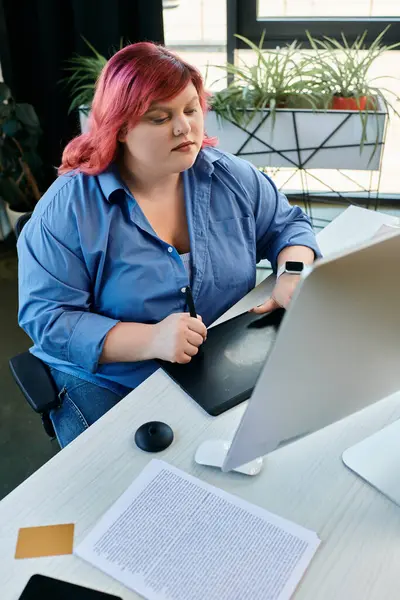 Una mujer de talla grande con cabello rosa usa una tableta de dibujo, enfocada en su trabajo. - foto de stock