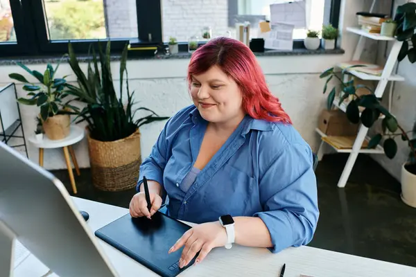 Una mujer de talla grande con el pelo rojo brillante dibuja en una tableta digital mientras trabaja en su escritorio. - foto de stock