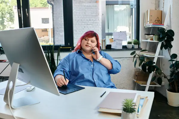 Una mujer de talla grande trabaja en una tableta gráfica, atendiendo una llamada telefónica en su escritorio. - foto de stock