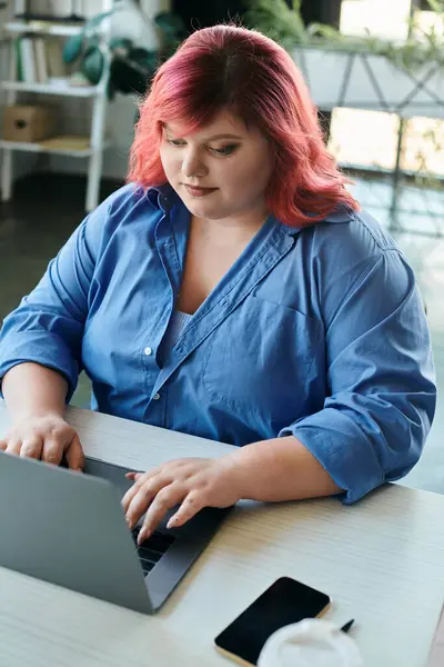 Женщина plus size с ярко-розовыми волосами сидит за письменным столом, работая над своим ноутбуком. — стоковое фото