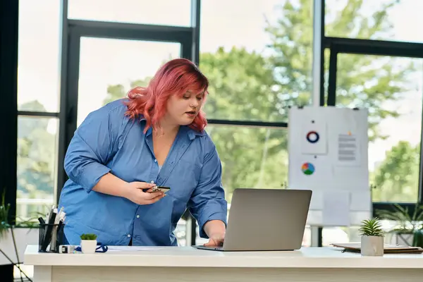 A plus size woman in a blue shirt uses a laptop while standing at a desk. — Stock Photo