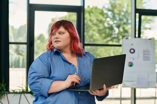 Женщина plus size в синей рубашке держит ноутбук и смотрит в сторону в офисной обстановке. — стоковое фото