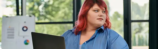 A plus size woman in a blue shirt sits at a desk, looking away from the laptop in front of her. — Stock Photo