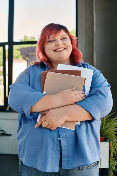 A plus size woman with bright red hair smiles warmly while holding a stack of books to her chest. — Stock Photo