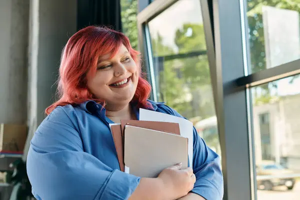 A plus size woman in a blue shirt, with vibrant red hair, smiles while holding books in her arms. — Stock Photo