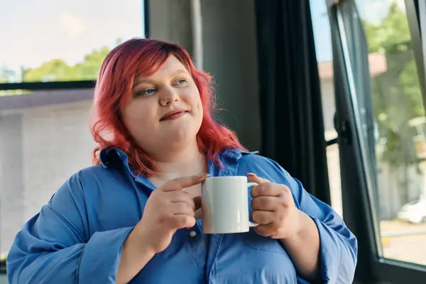 A plus size woman in a blue shirt contemplates, holding a white mug. — Stock Photo