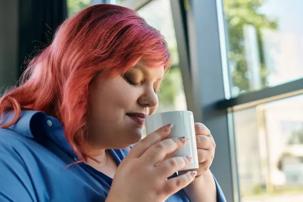 A plus size woman with pink hair sits by a window, enjoying a cup of tea or coffee. — Stock Photo