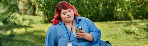 Una mujer de talla grande con una camisa azul y un pelo rojo vibrante se sienta en un césped verde y revisa su teléfono. - foto de stock