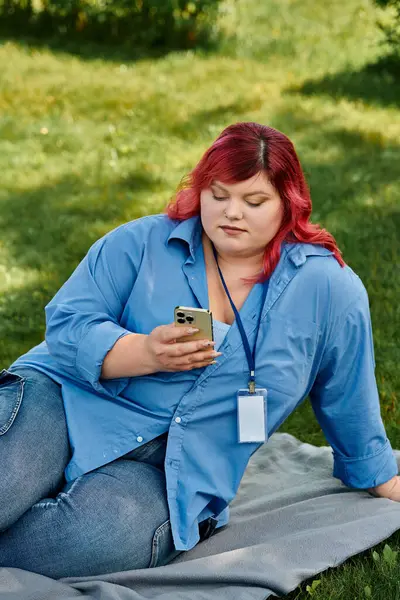 A plus size woman in a blue shirt and jeans sits on a blanket, scrolling on her phone. — Stock Photo