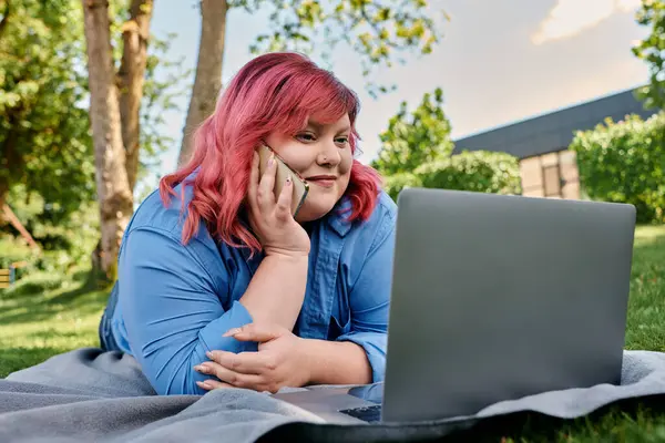 Una mujer de talla grande con el pelo rosa está hablando en su teléfono mientras está acostada en una manta en el parque. - foto de stock