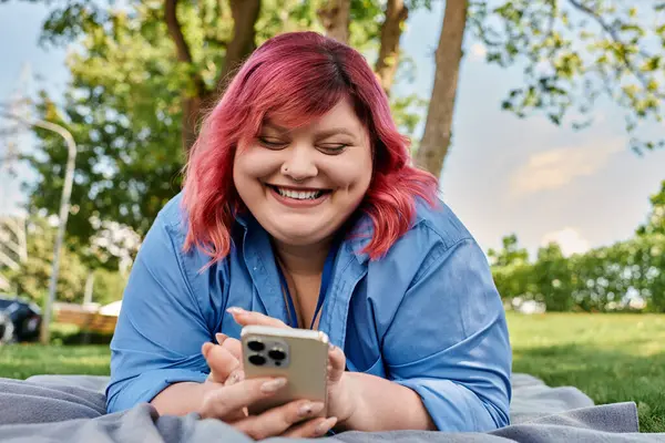 A plus size woman with pink hair lies on a blanket in a park, smiling at her phone. — Stock Photo