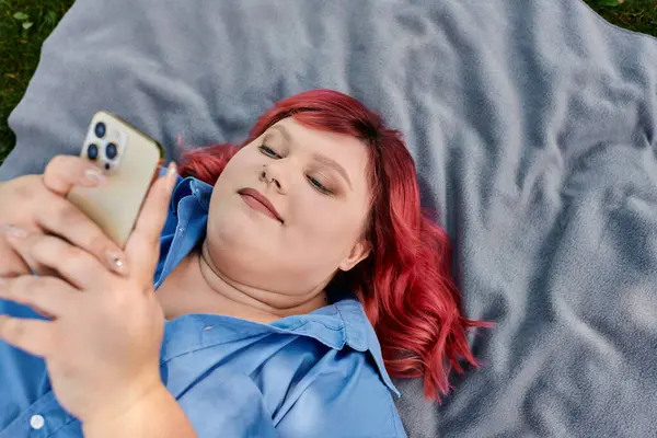 A plus size woman with red hair and a blue shirt lays on a gray blanket, scrolling through her phone. — Stock Photo