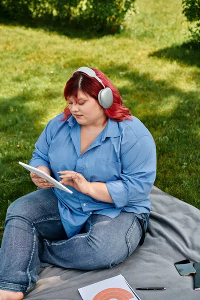 A plus size woman sits on a blanket in the grass, wearing headphones and browsing a tablet. — Stock Photo