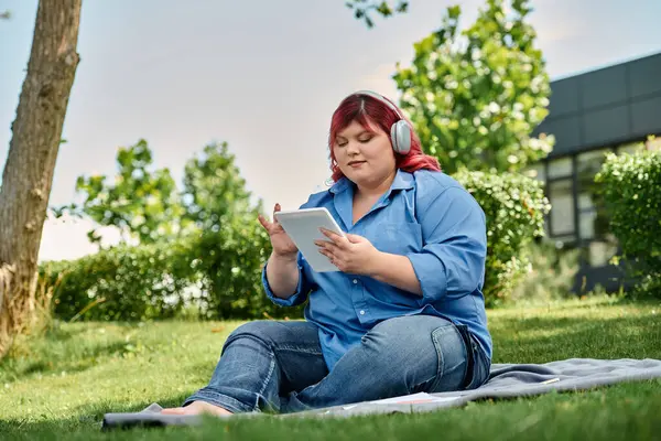 A plus size woman in blue and red attire uses a tablet while sitting on a blanket in a grassy park. — Stock Photo