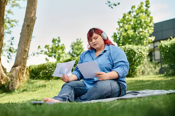 Женщина plus size в голубой рубашке сидит на одеяле, читая газеты в пышном зеленом саду. — стоковое фото