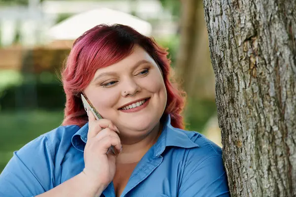 A plus size woman with bright pink hair speaks on her phone while standing by a tree. — Stock Photo