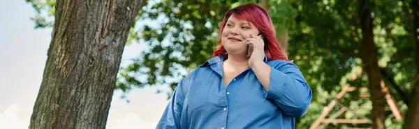 Eine Plus-Size-Frau mit roten Haaren geht durch einen Park, während sie mit ihrem Handy chattet. — Stockfoto