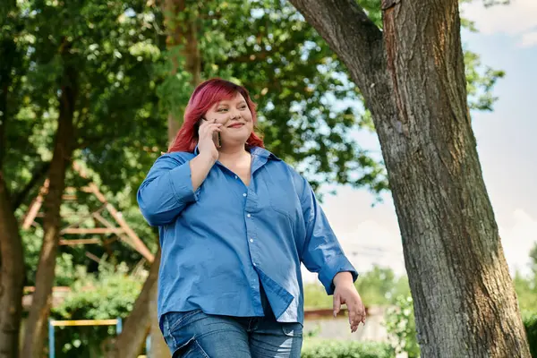 A plus size woman with bright red hair walks through a park while talking on her phone. — Stock Photo