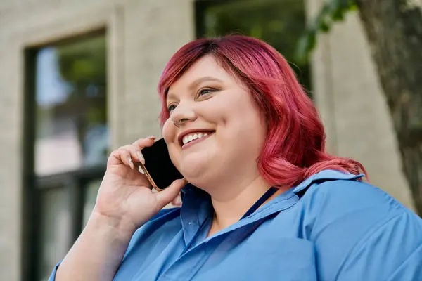 A plus size woman in a blue shirt talks on her phone while smiling brightly. — Stock Photo