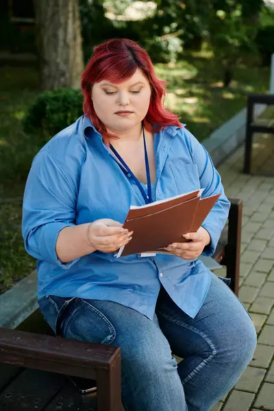 Eine Plus-Size-Frau mit roten Haaren sitzt draußen auf einer Bank und liest ein Buch. — Stockfoto