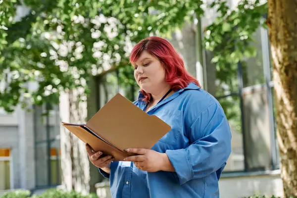 A plus size woman with bright red hair reads a folder outdoors. — Stock Photo