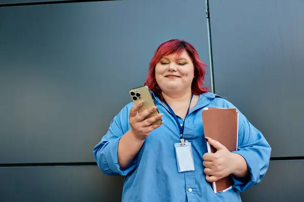 Eine Plus-Size-Frau mit leuchtend roten Haaren und blauem Hemd hält einen Ordner in der Hand und checkt lächelnd ihr Handy.. — Stockfoto