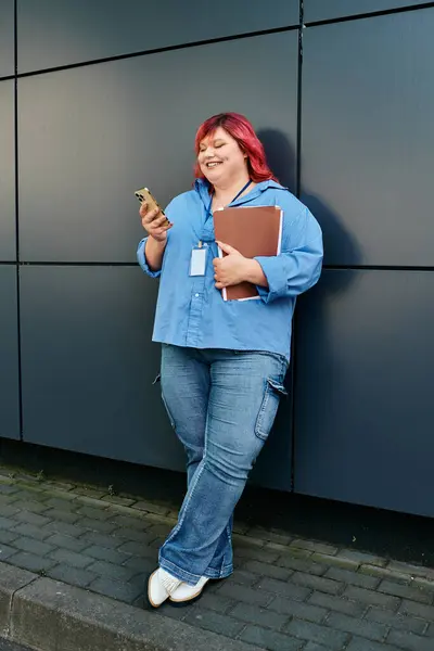 A plus size woman in blue leans against a wall, smiling and using her phone. — Stock Photo