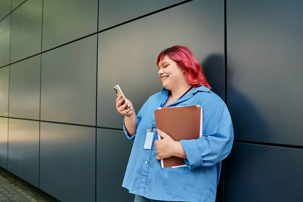 Uma mulher plus size em uma camisa azul com cabelo rosa verifica seu telefone enquanto segura uma pasta. — Fotografia de Stock