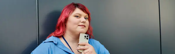 Una donna plus size con i capelli rossi luminosi posa con il suo telefono su uno sfondo grigio. — Foto stock