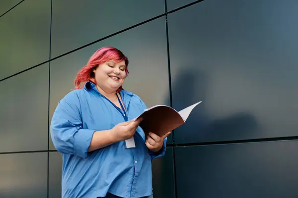 Une femme de taille plus dans une chemise bleue se dresse contre un mur d'acier, lisant une brochure. — Photo de stock