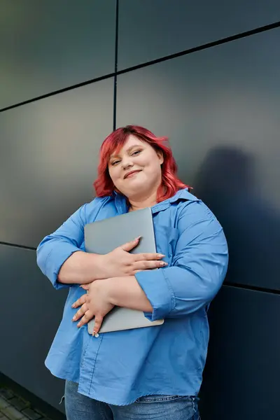 Red-haired plus size woman with confident smile holding laptop in front of a modern wall — Stock Photo