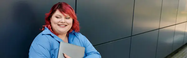 Smiling plus size woman with bright red hair holds laptop against grey wall — Stock Photo