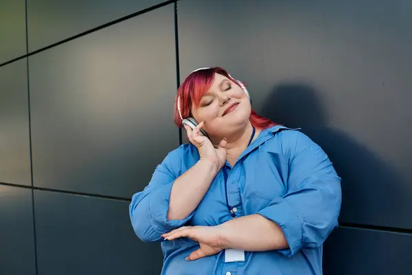 A plus size woman in a blue shirt and headphones leans against a gray wall, lost in the music. — Stock Photo