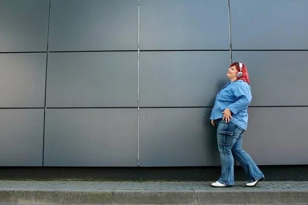 Una donna plus size in jeans blu e una camicia blu passa davanti a un muro grigio. — Foto stock