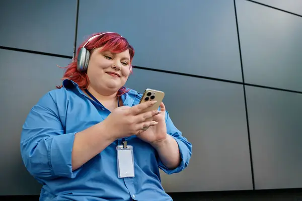Женщина plus size с розовыми волосами улыбается, когда она пользуется телефоном, в голубой рубашке и наушниках. — стоковое фото