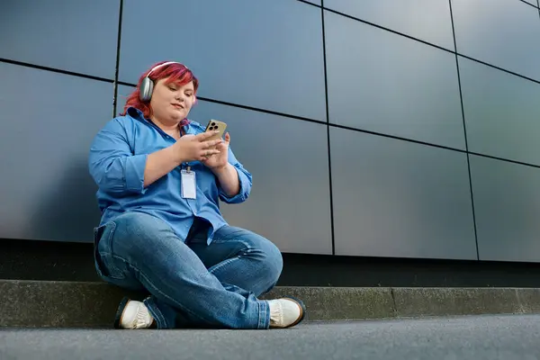 Una donna plus size si siede su un marciapiede di fronte a un edificio moderno, perso nel suo telefono. — Foto stock