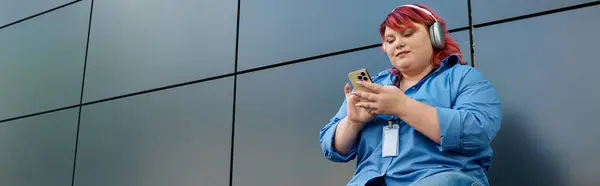 A plus size woman in a blue shirt and headphones looks at her phone against a tiled backdrop. — Stock Photo