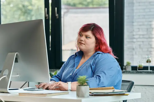 Una mujer de talla grande con cabello rosa se sienta en un escritorio trabajando en una computadora. - foto de stock