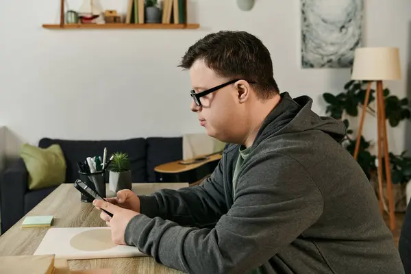 A young man with Down syndrome is focused on his phone while seated at his dining table. — Stock Photo